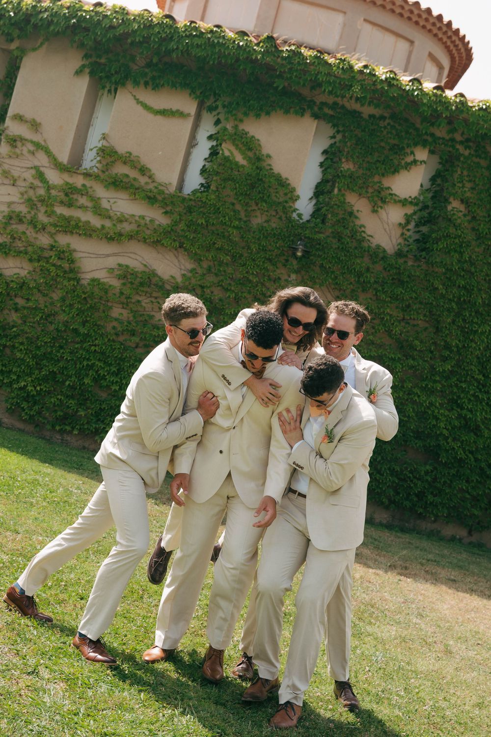 Groomsmen in cream-colored suits pose playfully on a lawn with ivy-covered walls in the background.