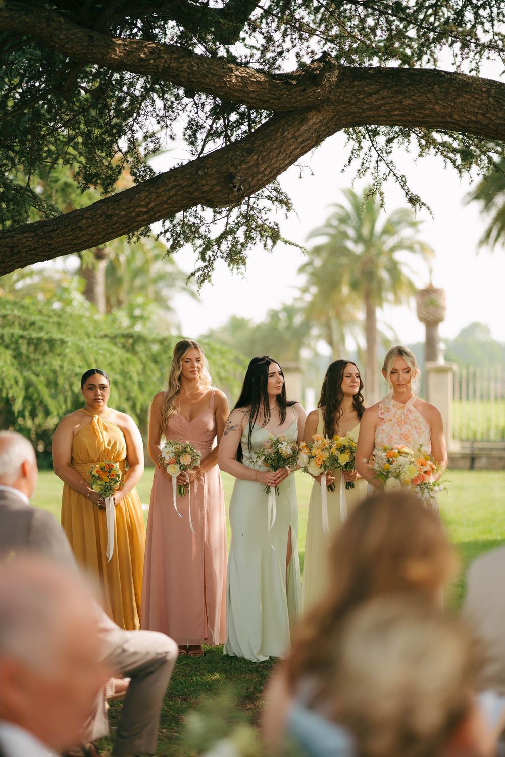 Des demoiselles d'honneur en robes fluides de jaune, rose et vert menthe se tiennent en ligne sous un arbre lors d'une cérémonie de mariage