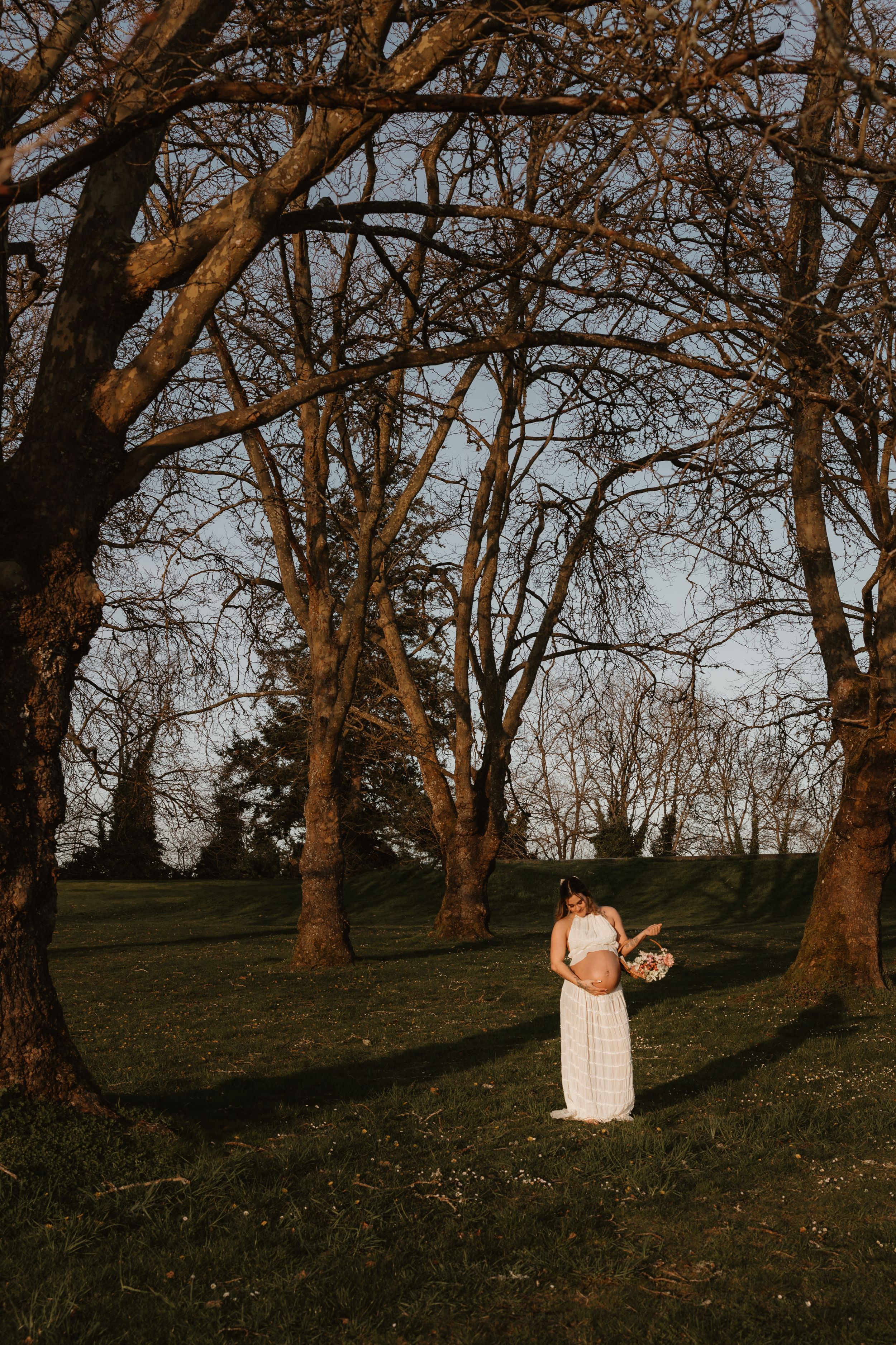A figure in a flowing white dress stands among tall bare trees at sunset in a serene outdoor setting.