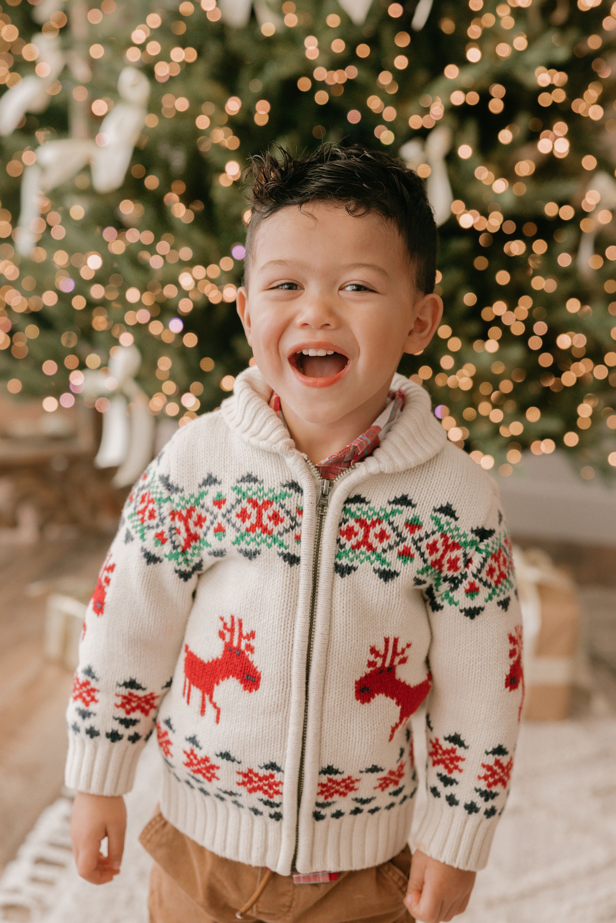 A joyful child wearing a festive white knit sweater with red reindeer pattern stands in front of twinkling Christmas lights.