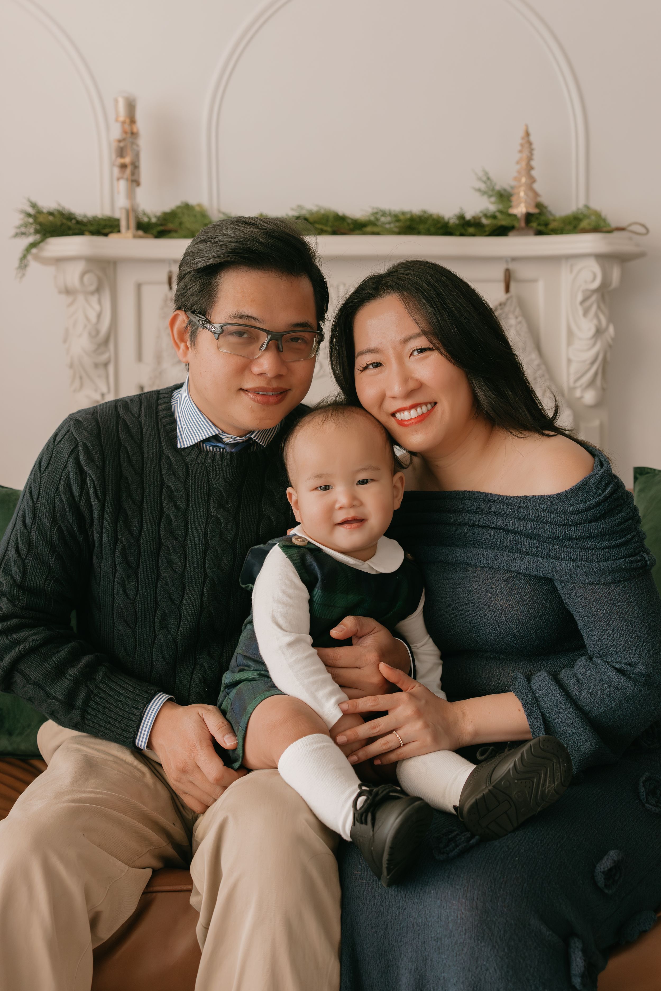 A young family poses together in front of a white fireplace mantel decorated with greenery for a holiday portrait.