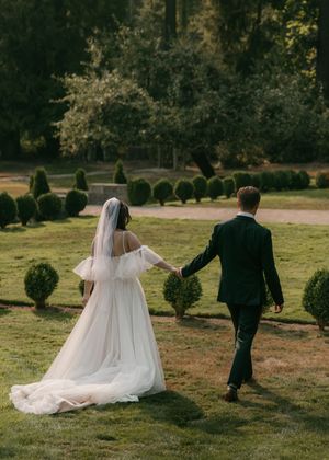 A couple walks hand in hand through a garden with manicured topiaries on their wedding day, the bride in a flowing white gown.