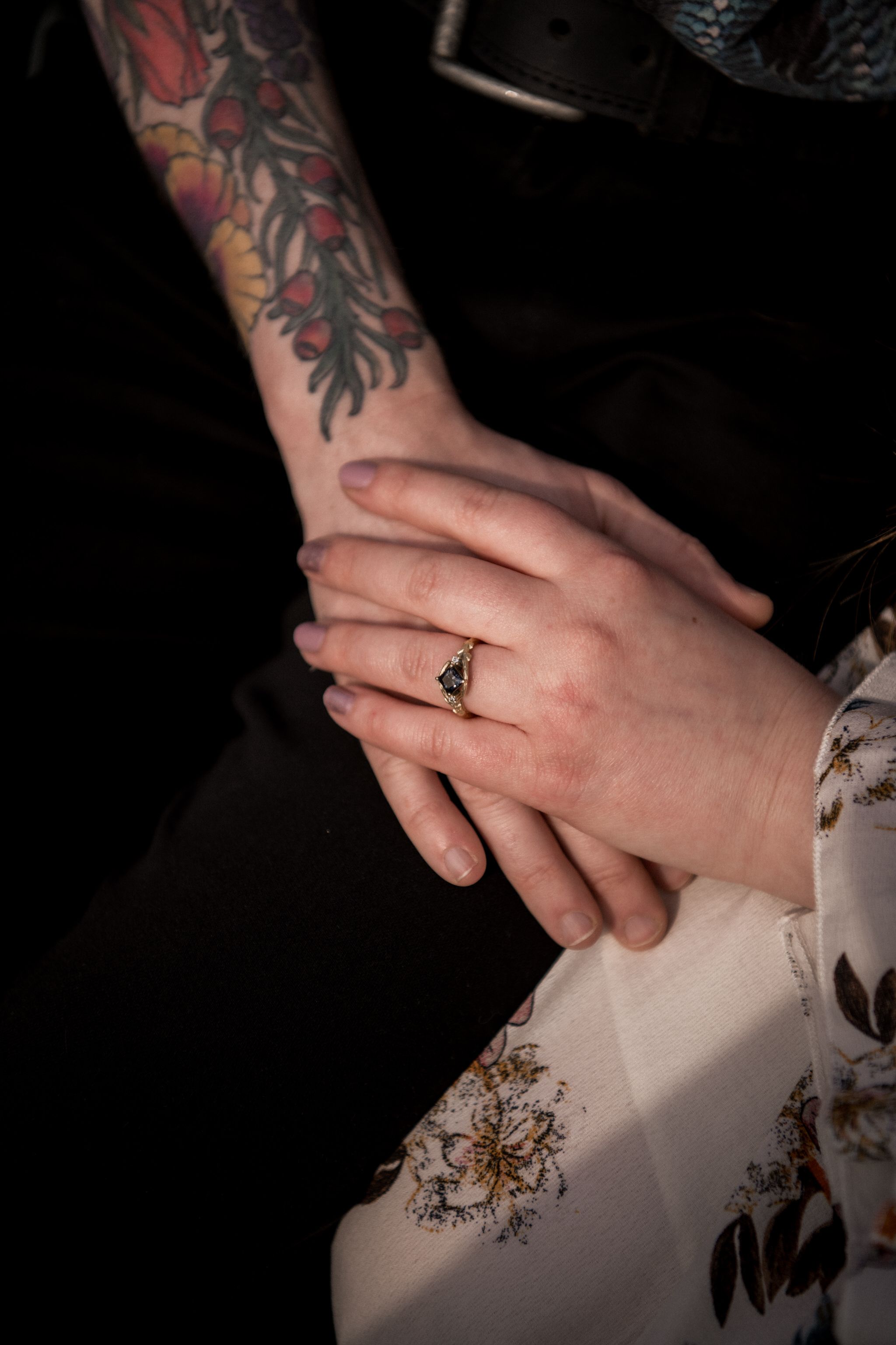 Two hands clasped together showing a gold ring and colorful floral tattoo sleeve against a dark background.