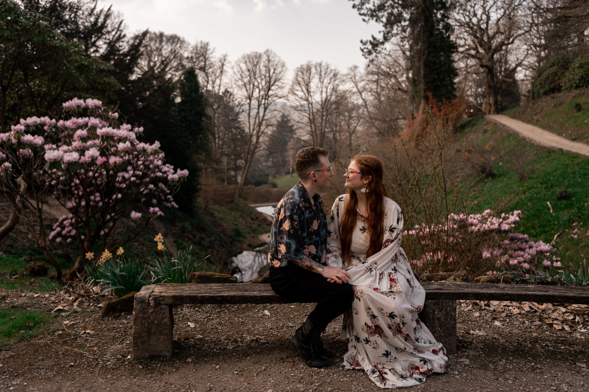Couple sits on stone wall surrounded by blooming cherry blossoms in a romantic spring garden setting.