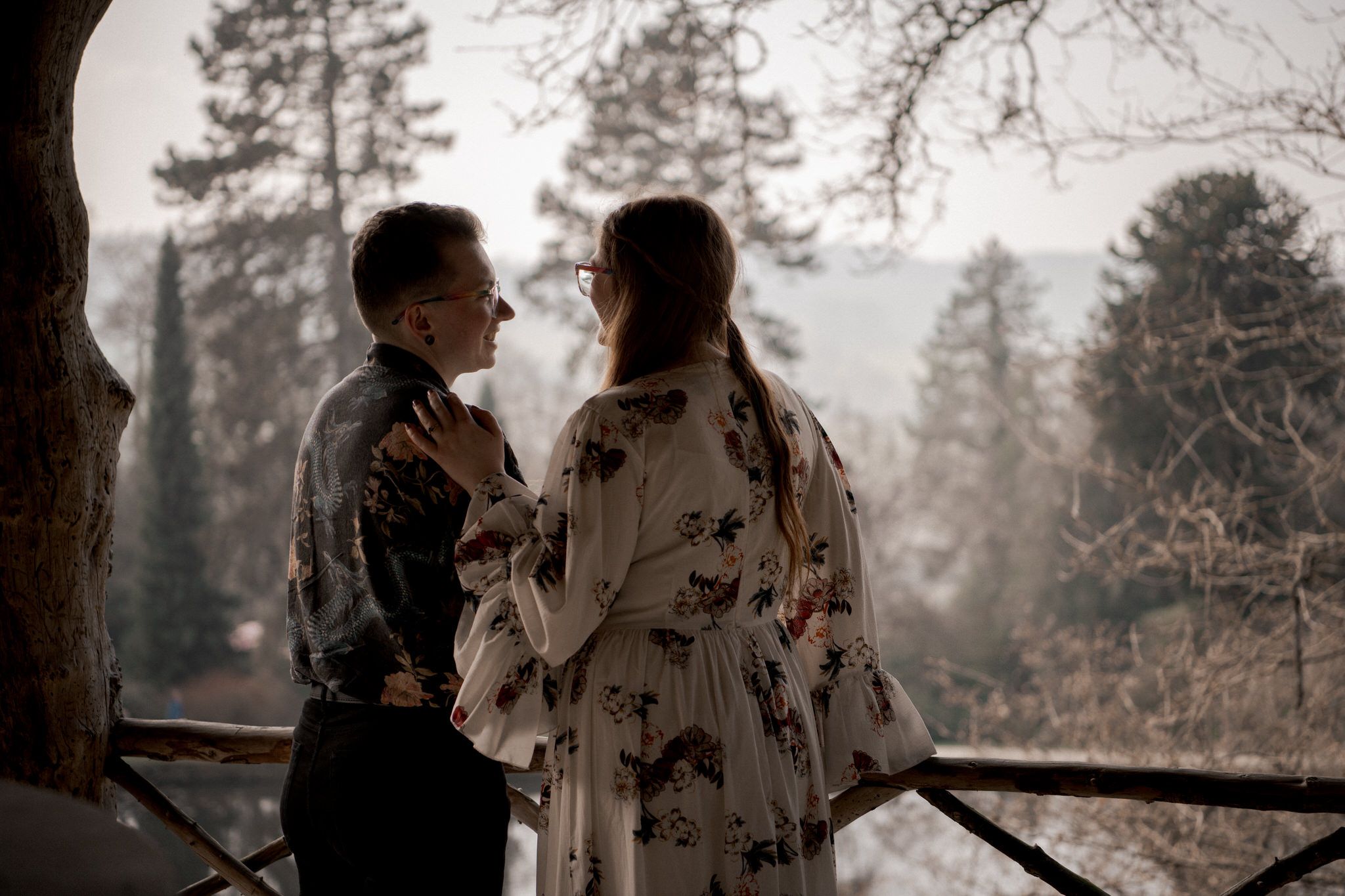A romantic couple stands on a misty forest balcony at dusk, silhouetted against evergreen trees in the background.