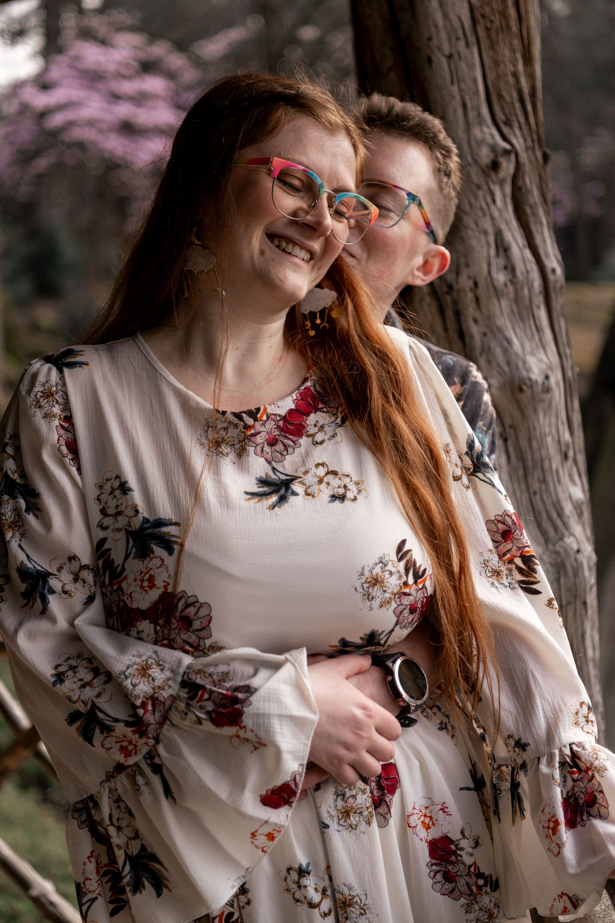 A couple wearing glasses share an intimate moment against a tree trunk with pink blossoms in the background.