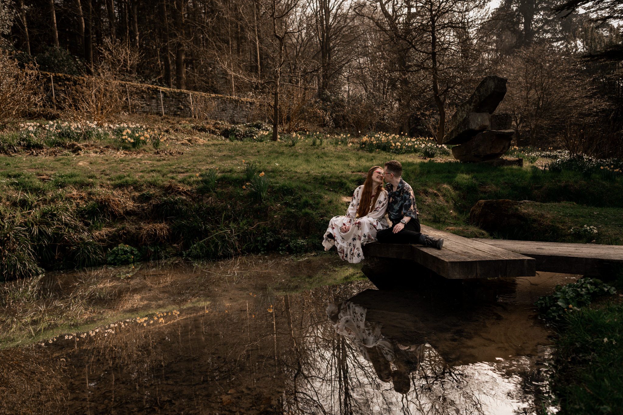A couple embraces on a rustic stone bridge surrounded by lush forest scenery in natural lighting.