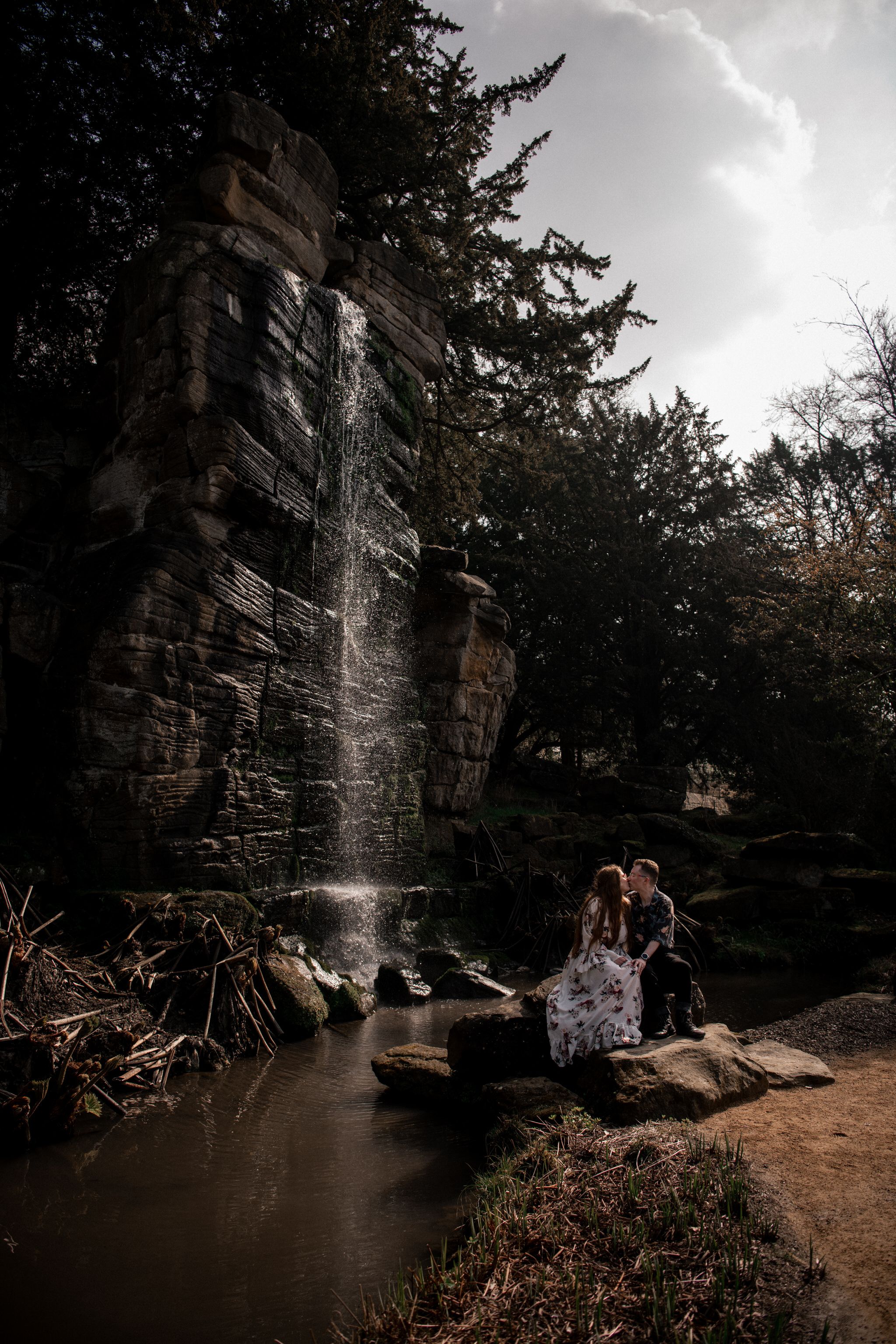 Dramatic waterfall cascades down rocky cliffs beside a tranquil pond in a moody natural setting.