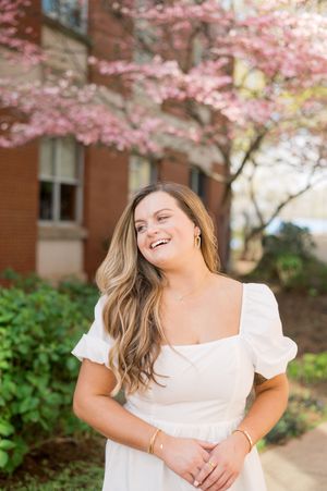 A person in a white dress smiles joyfully under blooming pink cherry blossoms near a brick building.