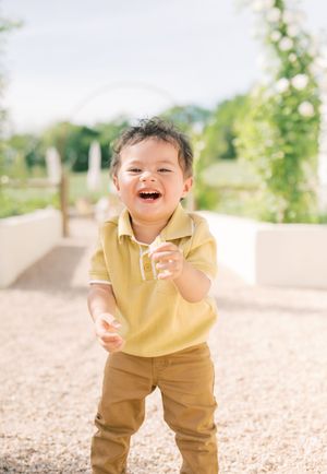 A child in a yellow polo shirt and brown pants laughs joyfully while playing outdoors on a sunny day.