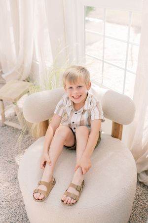 Child sits happily on a plush white chair by a bright window wearing sandals and summer clothes.