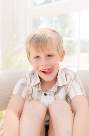 A young child wearing a light colored button-up shirt sits and smiles brightly in a sunlit room.