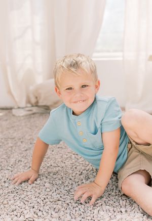 A toddler in a light blue shirt crawls and plays on a plush gray carpet in a sunlit room.