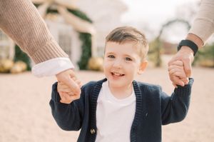 A small child wearing a white shirt walks while holding hands with adults on a sunny day.