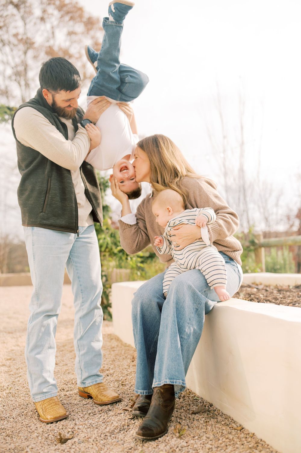 A family plays and laughs together outdoors on a sunny day, with a child being held upside down in a playful moment.