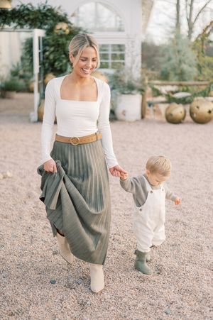 A parent and toddler in coordinating gray and white outfits walk together on a gravel path in an outdoor garden setting.
