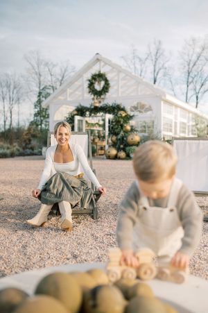 A rustic autumn scene outside a white farmhouse with a Christmas wreath, featuring a wooden wheelbarrow and fresh pumpkins.