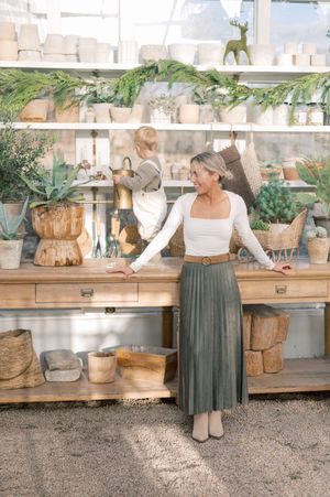 A person wearing a pleated sage green skirt and white top stands in a garden shop surrounded by plants and pottery.