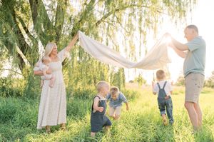 Family wearing white and blue outfits poses together in a sunlit meadow with willow trees at sunset.