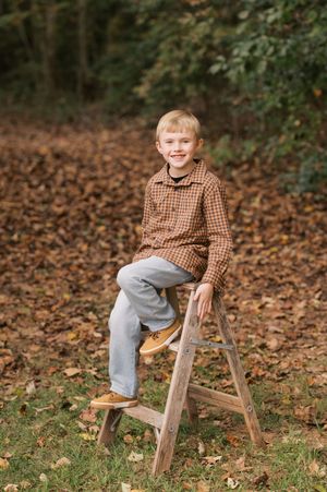 A child in a plaid shirt and gray pants sits on a wooden ladder surrounded by autumn leaves in an outdoor setting.