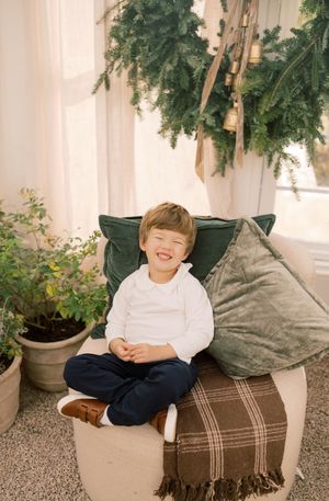 A child sits on a brown plaid blanket outdoors with potted plants and white columns in the background.