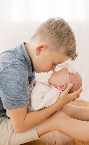 A young boy in a blue shirt gently kisses a newborn baby lying on a white surface in a bright room.