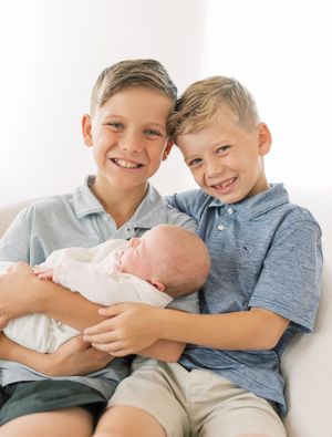 Three siblings sitting together on a white background sharing tender moments with their newborn baby sibling.