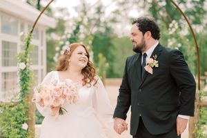 Joyful wedding couple laughs together under a floral arch with pink bouquet.