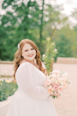 A bride in a flowing white veil holds a romantic bouquet of pink roses and flowers in an outdoor garden setting.
