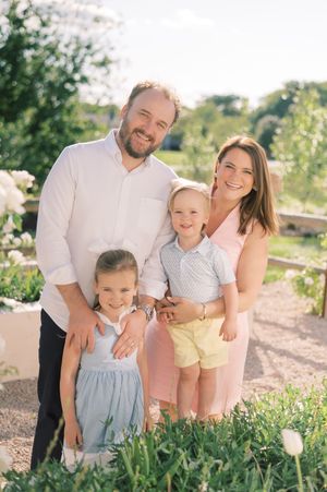 Family wearing coordinated white and pastel outfits pose together in a sunny outdoor garden setting.