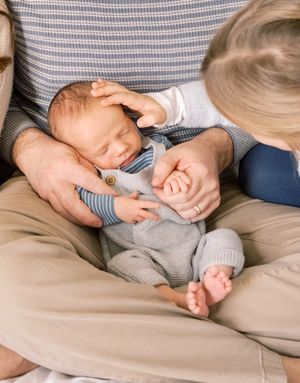 A sleeping infant in gray clothing rests on someone's lap while being gently touched by small hands.