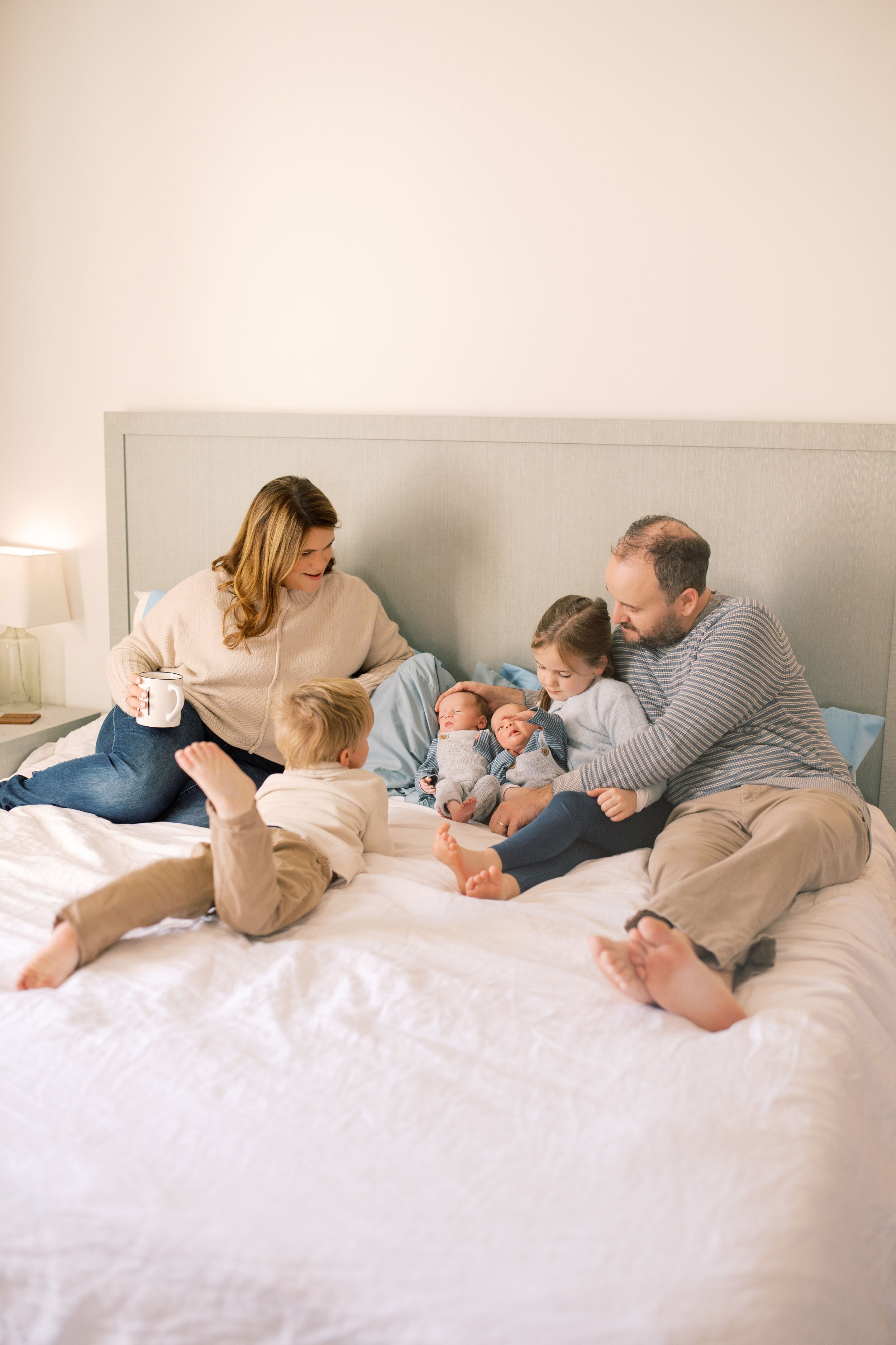 Family relaxing together on white bed with children and baby in casual clothing at home.