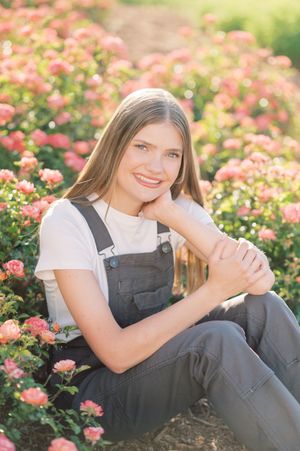 A person in gray overalls sits among blooming pink roses in a garden during a sunny day.