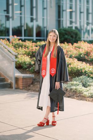 Graduate in black cap and gown with red stole stands outdoors near flower garden wearing red platform sandals.