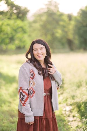 A woman in a brown dress and gray patterned cardigan poses outdoors in a sunlit field.