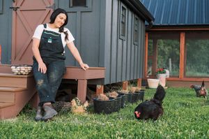 A person in overalls stands near chickens on the grass outside a modern tiny house with dark siding.