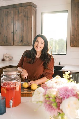 Someone preparing food in a bright kitchen with wooden cabinets, pink flowers, and a glass dispenser of red beverage.