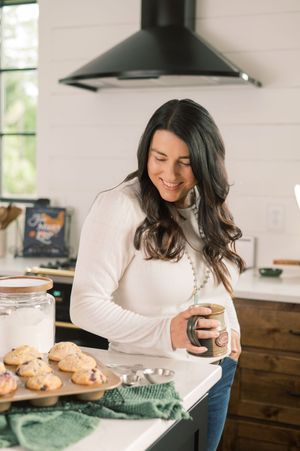 A woman in a white sweater smiles while baking cookies in a cozy kitchen with fresh baked goods on the counter.