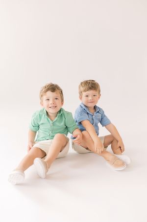 Two young children in collared shirts and shorts sit together on a white background for a studio portrait.