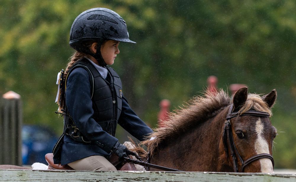 A young female equestrian wearing a helmet sits on a horse looking away from the camera while the horse looks toward the camera in the rain.