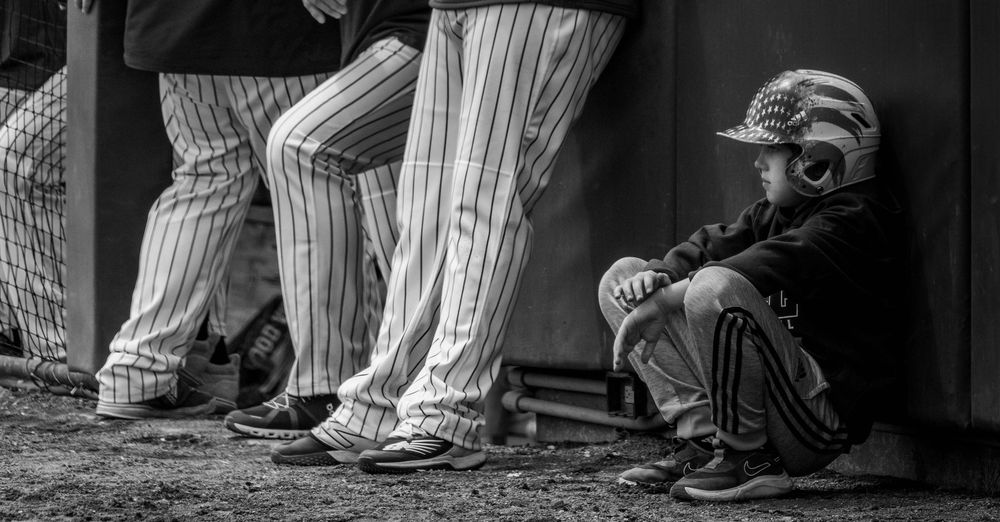 Black and white image of a boy wearing a batting helmet and sitting outside the dugout. Older baseball players stand next to him.