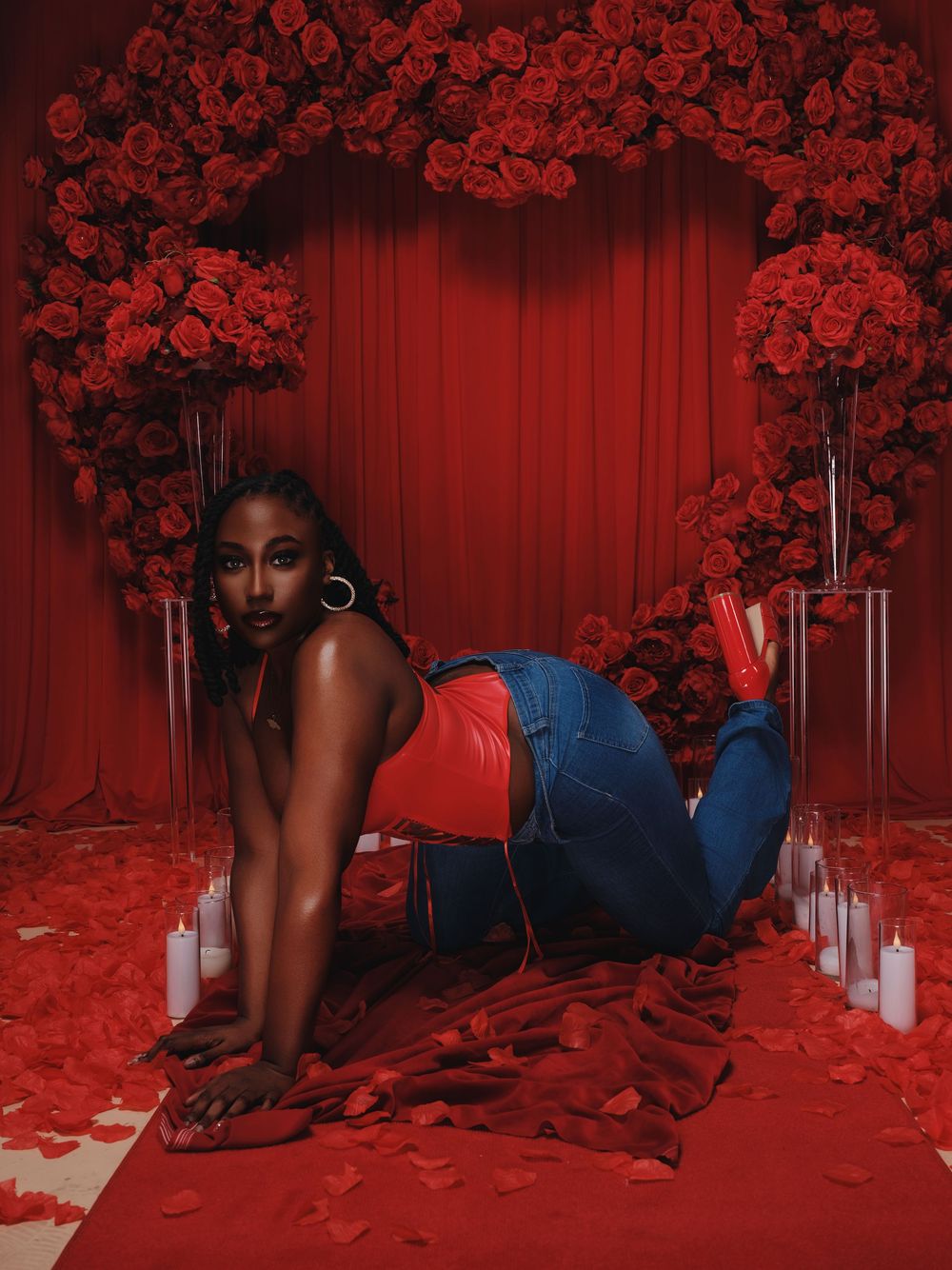 A dramatic red-tinted photoshoot setup with rose arrangements, candles, and rose petals scattered on the floor.