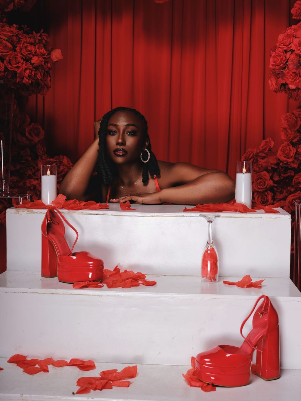 Artistic bathtub scene with red roses and candles against dramatic red curtain backdrop creates romantic atmosphere.