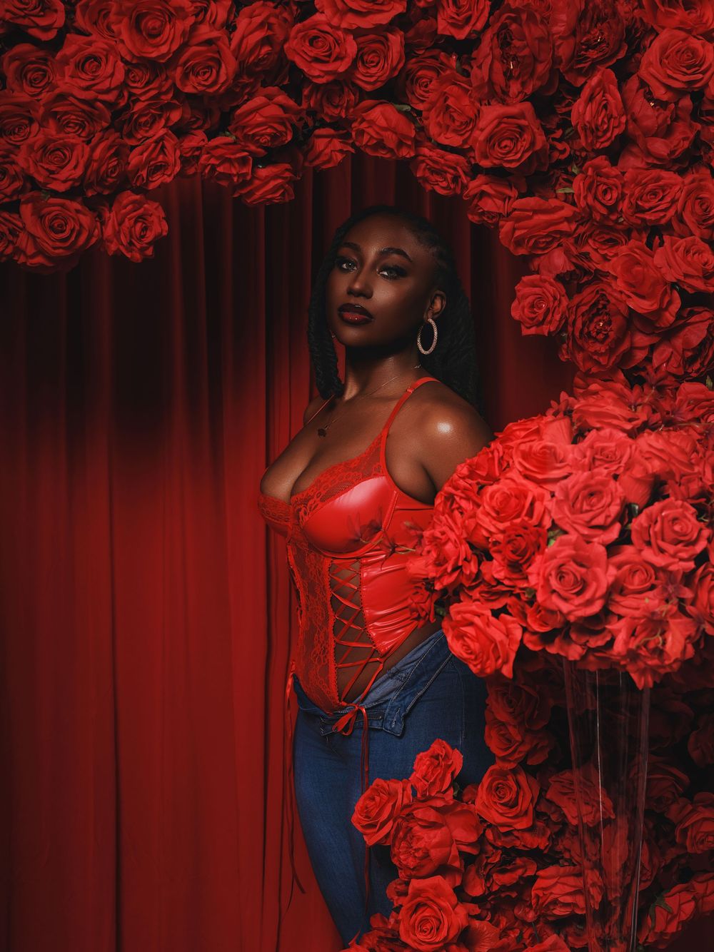 A dramatic portrait against a red curtain backdrop with vibrant red roses framing the composition in rich crimson tones.