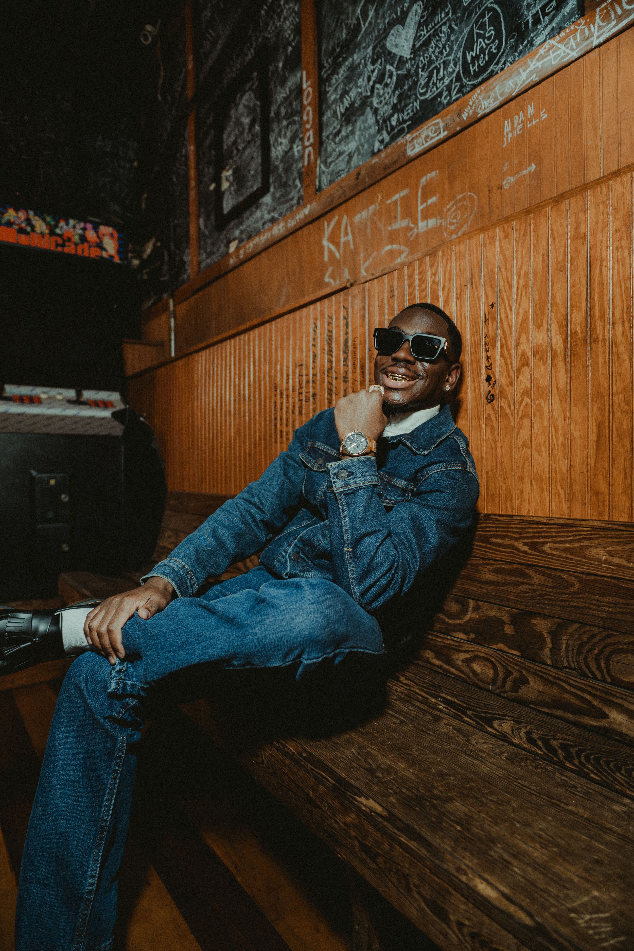 Person in denim outfit and sunglasses reclines against wooden wall in artistic photo series.