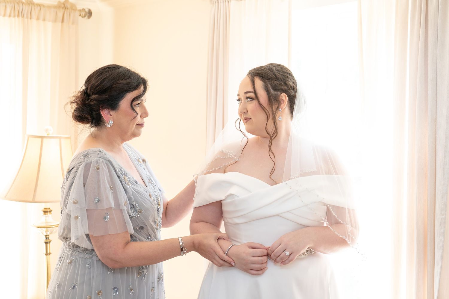 A bride in a white gown and veil being helped by a woman in a floral dress in a bright, elegant room.