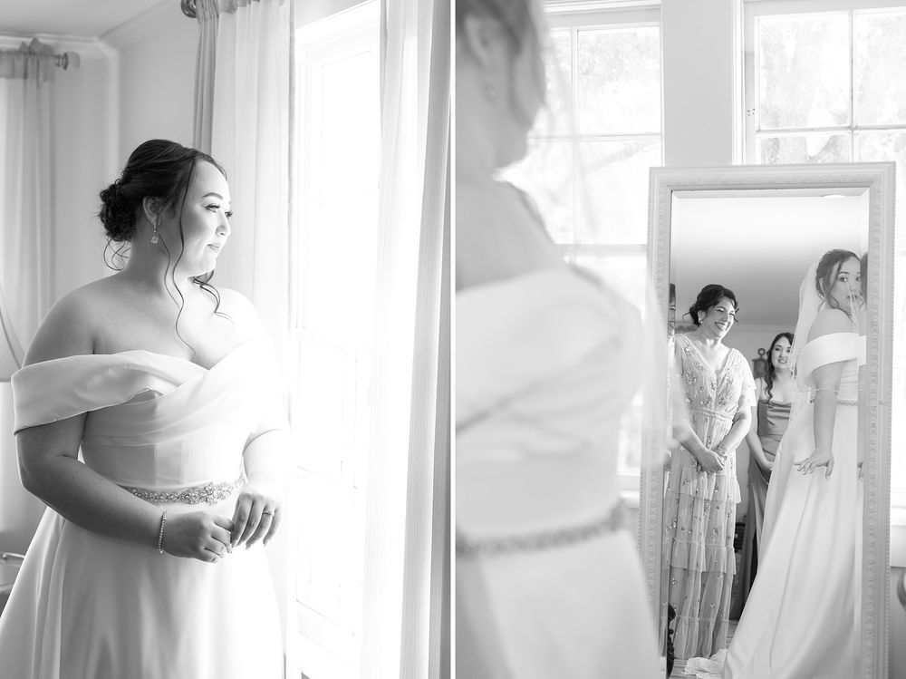 Black and white photo of a bride in an off-shoulder gown gazing out a window while attendants help her prepare.
