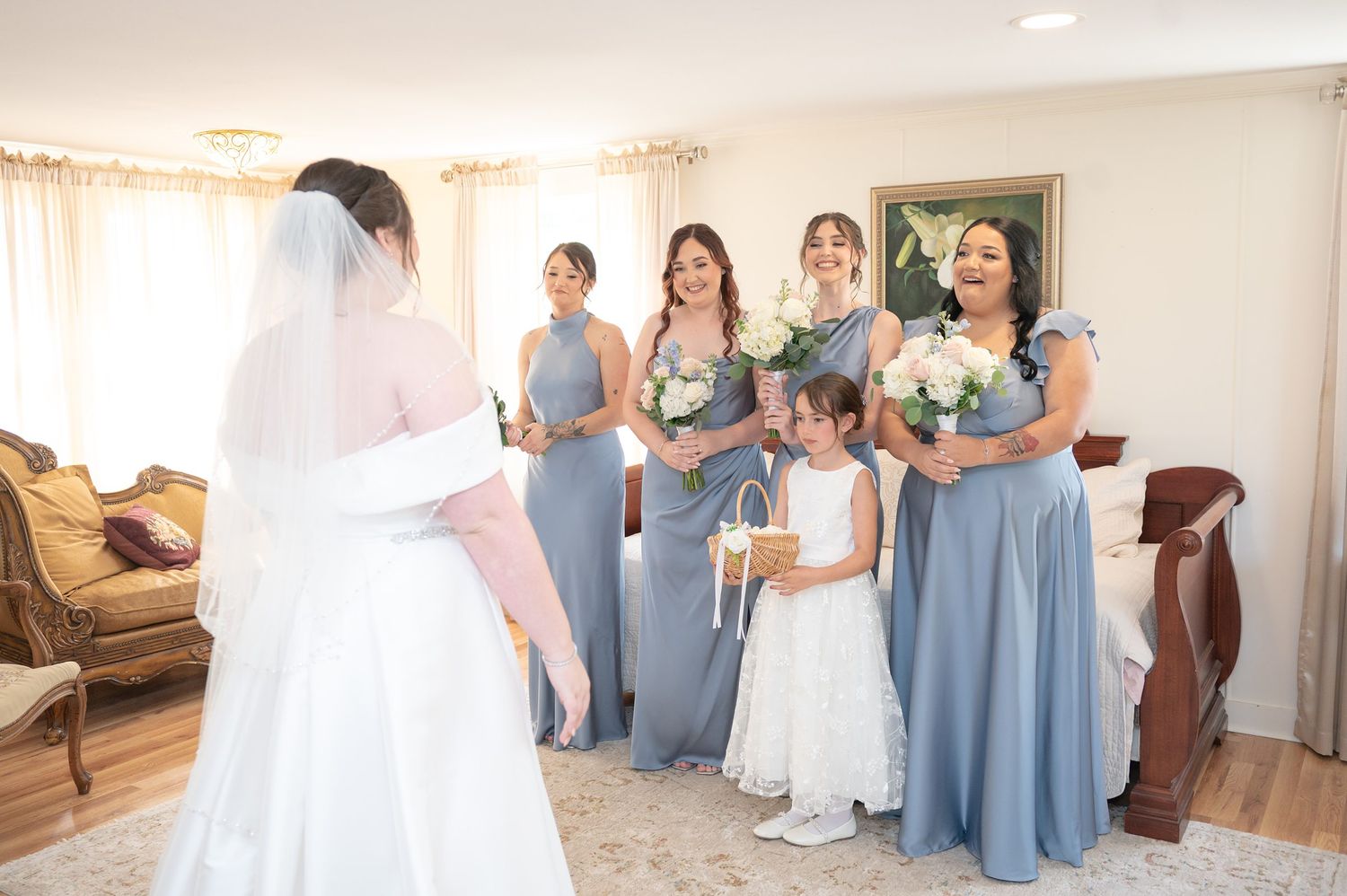 Bride in white gown and veil reveals herself to smiling bridesmaids in blue dresses and flower girl in white dress.