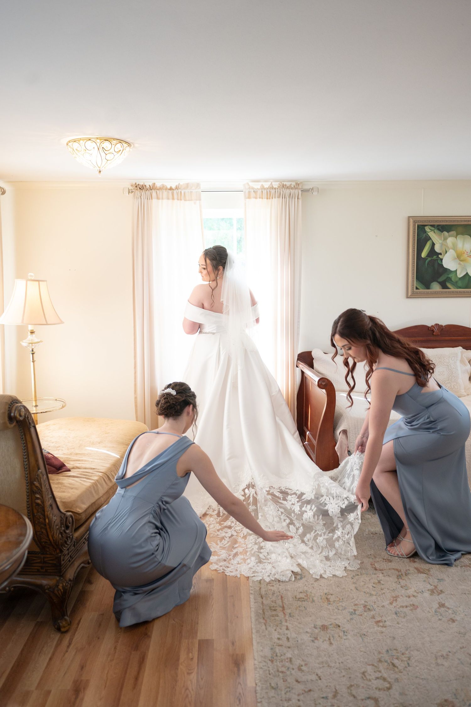 Bridesmaids in blue-gray dresses arrange the lace train of a bride in a white wedding gown standing by a sunlit window.
