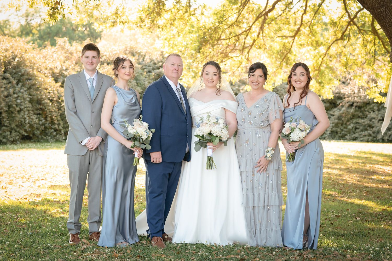 Bride in white gown poses with wedding party in gray dresses and suits outdoors under golden autumn trees.